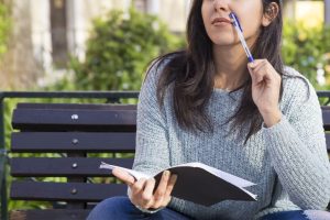 Woman sitting on park bench and pausing to think while writing in a notebook